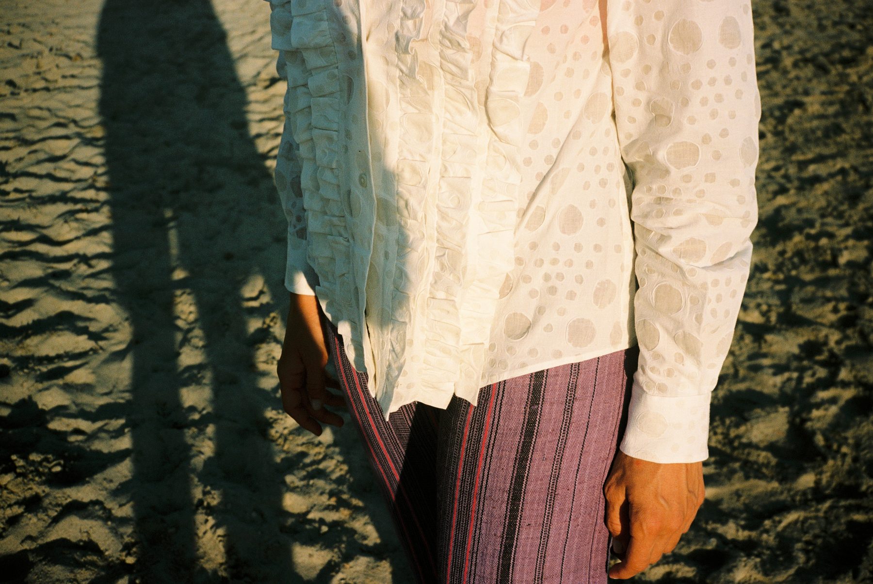 Scan 16 — white eyelet ruffle shirt and purple stripe trousers, long shadow on sand